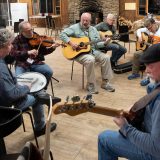 A group sitting in a circle playing a song. A woman playing the drums, a man playing the fiddle, another man playing the bass and four men playing the guitar