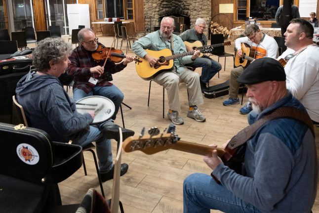 A group sitting in a circle playing a song. A woman playing the drums, a man playing the fiddle, another man playing the bass and four men playing the guitar